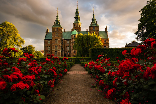 Brick castle with green copper spires framed by vibrant red roses along a garden pathway.