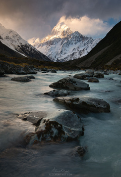 Snow-capped mountain peak rising above a glacial river with boulders in the foreground.