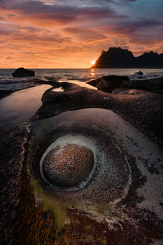 Rocky coastal landscape at sunset with circular stone formation in foreground and distant island silhouette.