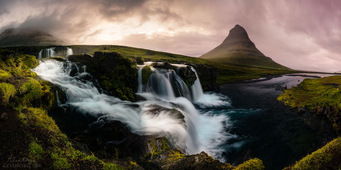 Cascading waterfall with moss-covered rocks flowing toward a distinctive pyramidal mountain peak under dramatic cloudy sky.