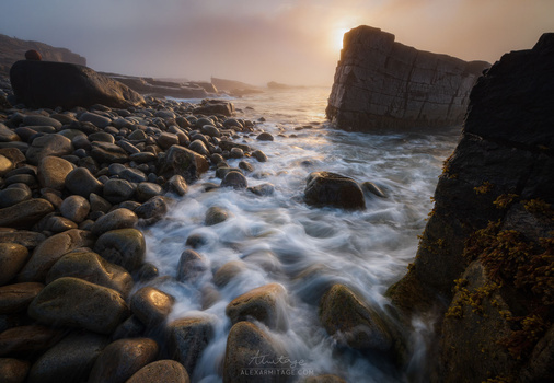 Rocky coastline at sunset with smooth ocean waves flowing around boulders and sea stacks.