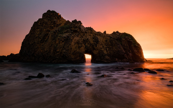 Large rocky sea arch silhouetted against golden sunset with waves washing across beach.