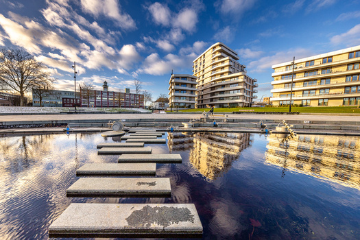 Concrete stepping stones reflect in still water with modern apartment buildings and cloudy sky beyond.