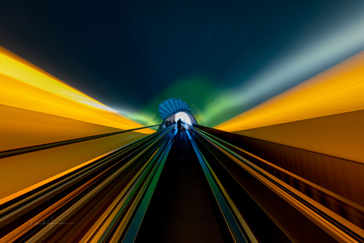 Long exposure view down a modern tunnel with symmetrical yellow-orange walls, railroad tracks converging toward a bright vanishing point, and teal-green light streaming from above.