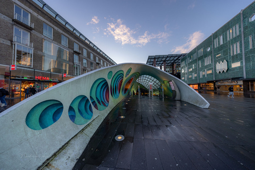 Curved sculptural archway with turquoise circular cutouts in an urban plaza surrounded by buildings.