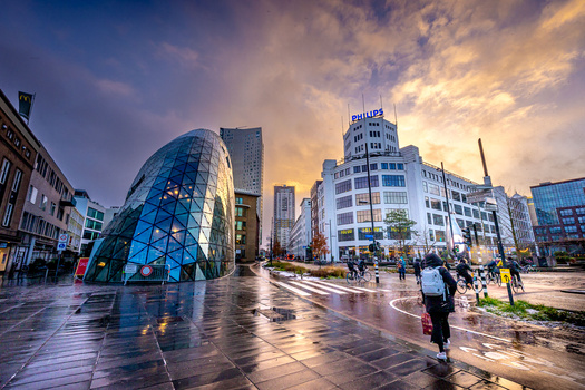 Urban plaza at dusk with wet pavement, modern curved glass building, and city skyline under golden-hour clouds.