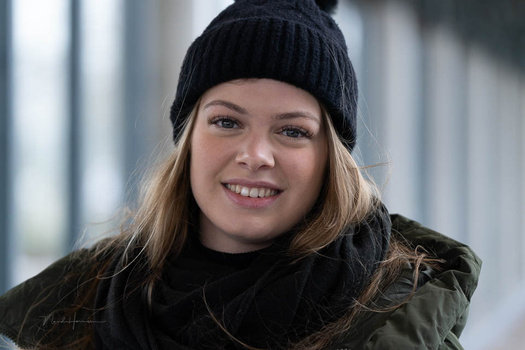 Young woman with blonde hair wearing a black beanie and olive jacket, smiling at the camera outdoors.