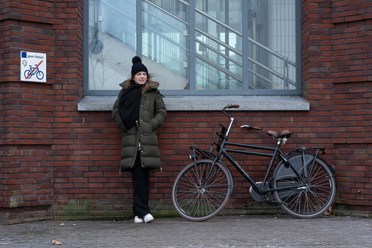 Woman in olive puffer jacket standing against red brick wall with two bicycles beside her.