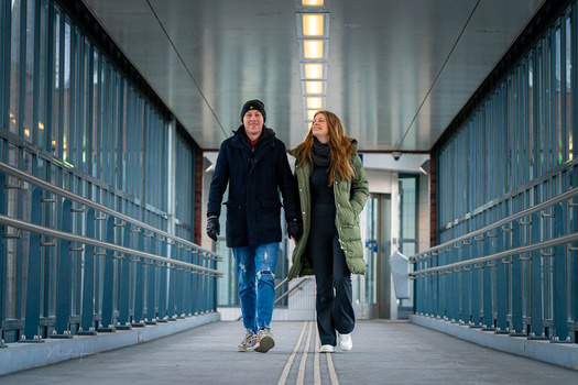 Two people walking toward the camera down a symmetrical corridor lined with metal gates and overhead fluorescent lighting.