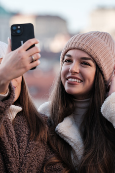 Two smiling women taking a selfie outdoors on an overcast day, one wearing a beige knit beanie.