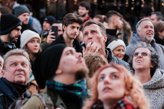 Diverse crowd of people gathered outdoors at dusk, gazing upward with expressions of wonder and anticipation.