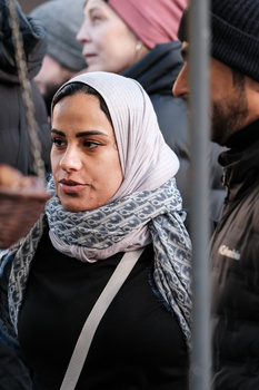 Woman wearing a hijab and gray scarf looking toward the camera amid a crowd.