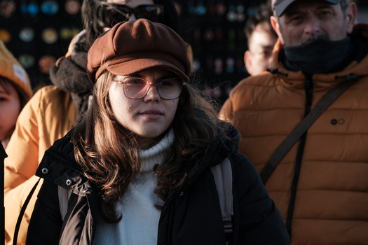 Person wearing brown cap, glasses, and layered winter clothing stands among a crowd outdoors.