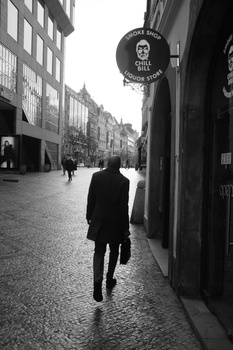 Person in dark coat walking away down a cobblestone street lined with historic buildings in black and white.