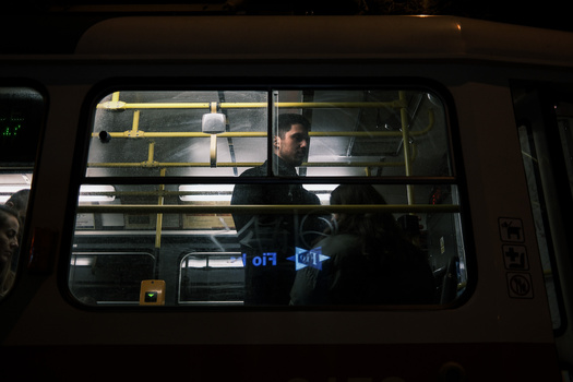Man standing inside a public bus at night, viewed through the window with yellow handrails visible.