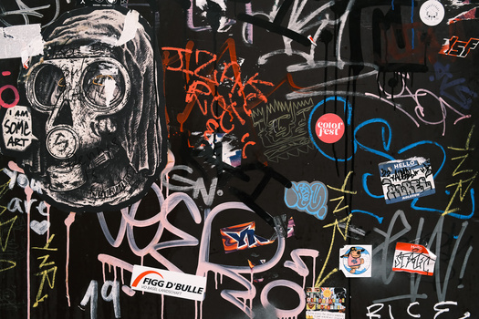 Dense graffiti wall covered in overlapping tags, throws, and character art in pink, blue, yellow, and white paint on black background.