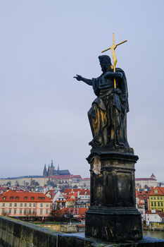 Dark bronze statue on a bridge pedestal with golden cross, overlooking a European city with red-roofed buildings and cathedral spires.