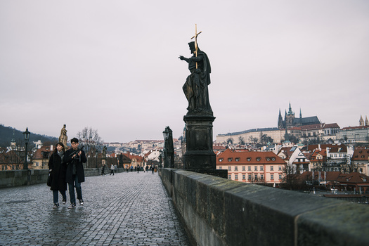 Statue of a robed figure on a bridge overlooking a European city with cathedral spires in the background.