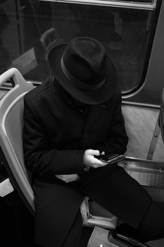 Person in dark coat and fedora hat sitting in transit, looking down at mobile device.