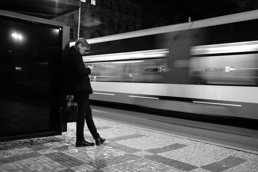 Man in dark coat standing on platform as blurred train passes by at night.