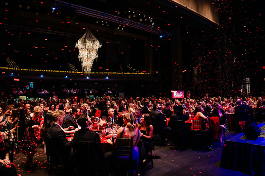 Large ballroom filled with formally dressed guests seated at illuminated tables beneath a crystal chandelier.