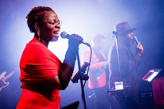 Female singer performing at microphone in red outfit with blue stage lighting and backup performers behind her.
