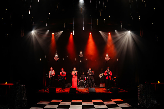 Band performing on stage under dramatic red and white lighting with checkered floor.