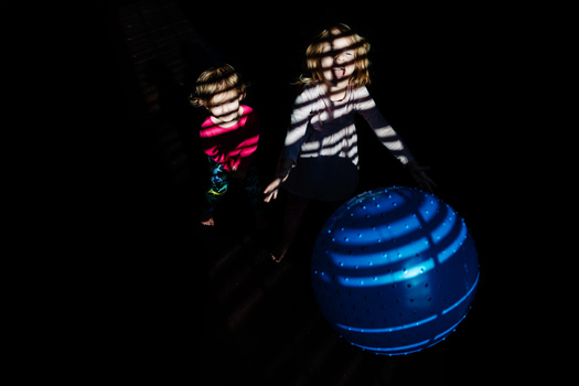Two children in striped shirts interact with a large illuminated blue sphere against a dark background.