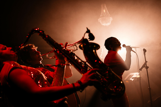Silhouetted musician singing at microphone with saxophonist and drummer visible in warm stage lighting.