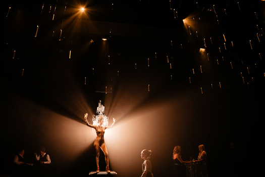 Theatrical performer suspended in dramatic spotlight with sparks falling overhead while ensemble cast stands positioned around the stage.