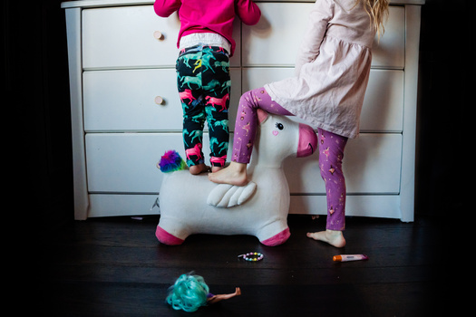 Two children in colorful clothing playing with a white toy unicorn on a dark floor in front of a white dresser.