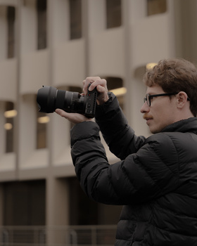 Photographer in black jacket holding DSLR camera with telephoto lens to his face, wearing glasses, shooting outdoors with blurred building in background.