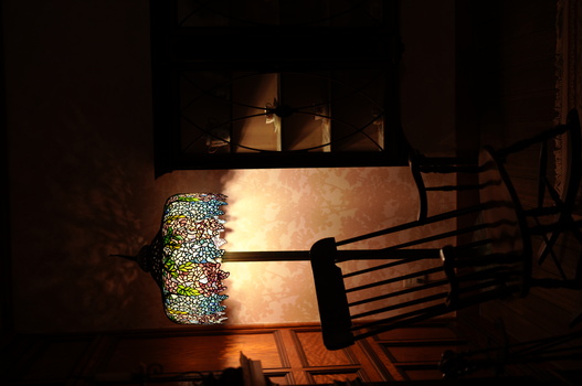 Tiffany-style stained glass floor lamp with green and amber glass dome illuminating a wooden rocking chair in a dimly lit interior space.