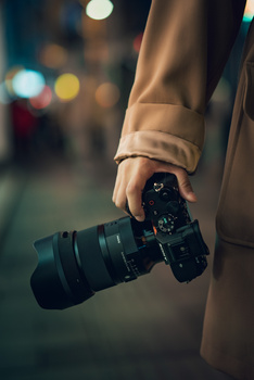 Photographer holding a camera with telephoto lens at night on an urban street with blurred city lights.
