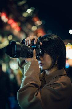 Young photographer holding a camera to their eye against a blurred nighttime cityscape backdrop.