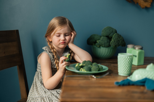 Young child at wooden table with plate of broccoli, resting chin on hand with reluctant expression.