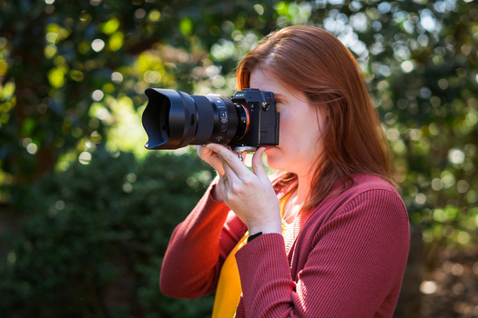 Female photographer with reddish-brown hair holding a mirrorless camera with telephoto lens to her face, wearing a burgundy cardigan outdoors.