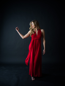Woman in flowing red dress against dark background with hand raised.