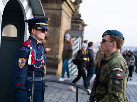 Military officer in dress uniform conversing with soldier in camouflage at a historic stone building.