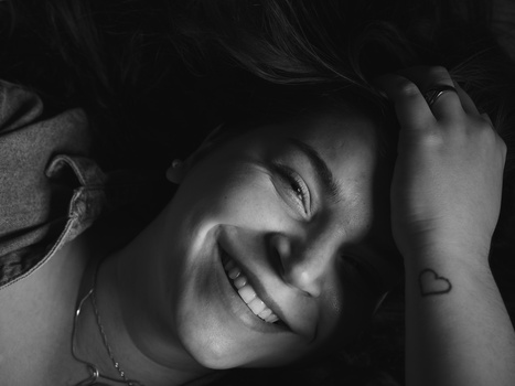 Black and white portrait of a smiling person with hand raised to head, shot from above at an intimate angle.