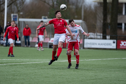 Soccer player in red jersey heads the ball during a competitive match while an opponent in white looks on.