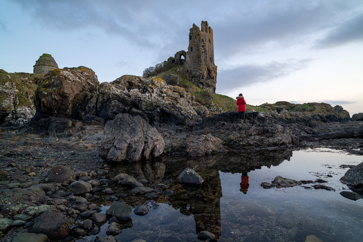 Ruined stone tower on rocky coastal outcrop with figure in red jacket reflected in tidal pool.
