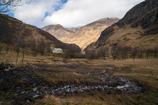 Stream flowing through a valley with mountains, bare trees, and a white house in the distance.