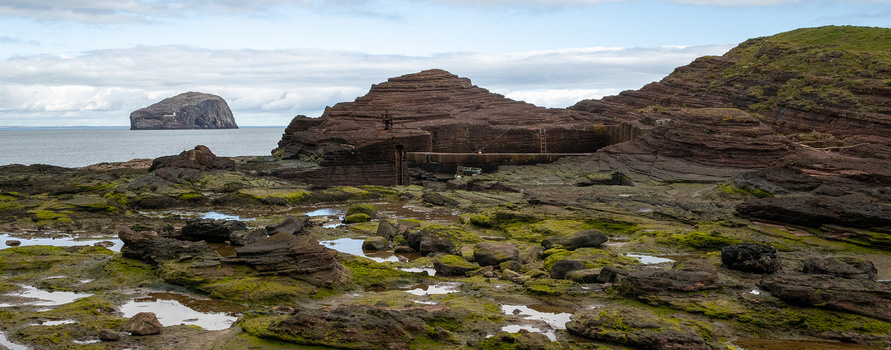 Rocky coastal landscape with moss-covered boulders, tidal pools, and a distant sea stack under partly cloudy skies.