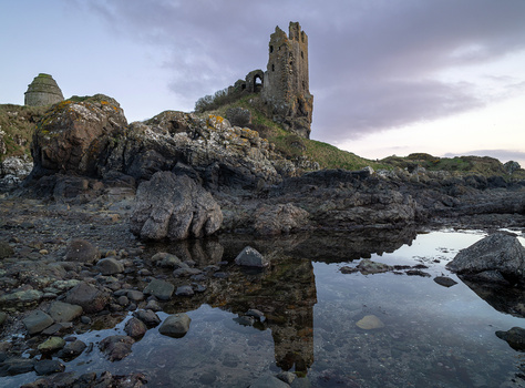 Ruined stone tower on rocky coastal outcrop reflected in tidal pool at dusk.