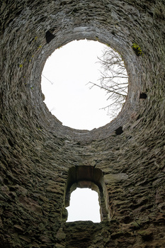 Upward view through two circular stone archway openings stacked vertically, framing bare tree branches and bright sky.