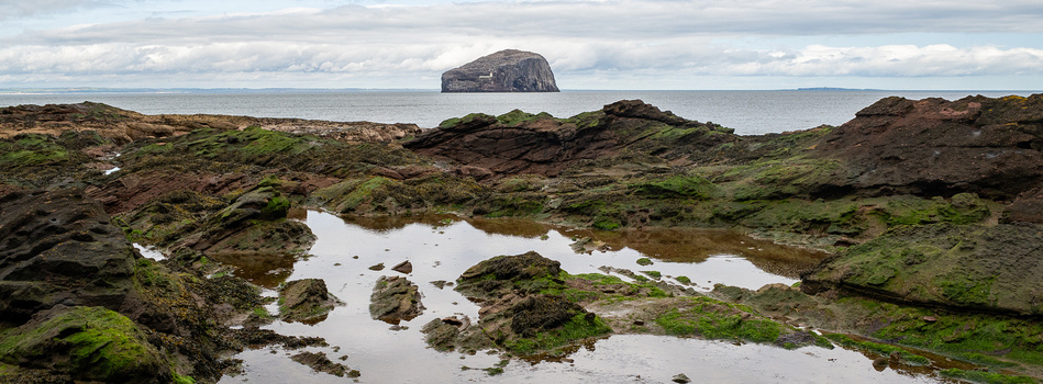 Rocky tidal pools with moss-covered boulders in foreground; distant island visible across calm sea under cloudy sky.