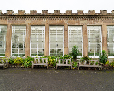 Historic stone building facade with tall mullioned windows and crenellated roofline, flanked by ornamental benches and potted plants.