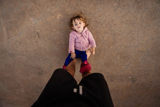 Toddler standing on adult's legs against textured brown background, shot from above.