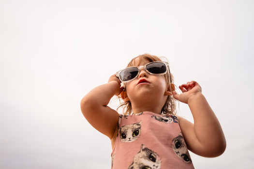 Toddler wearing oversized sunglasses and pink animal-print shirt, looking upward with hand near head.